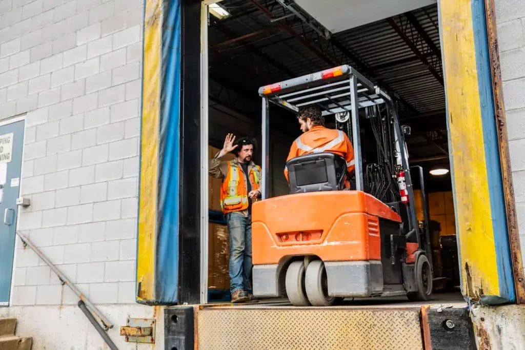 An industrial warehouse workplace safety topic. A manager or supervisor stops a forklift driver from backing up over a loading dock.